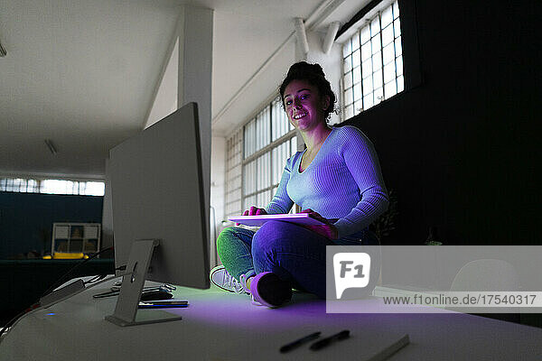 Young woman sitting on desk in front of desktops at home office