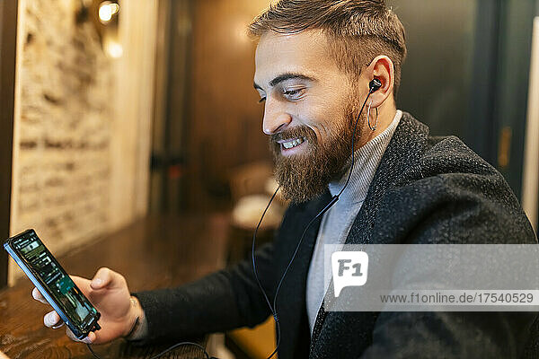 Glücklicher junger Mann  der sein Mobiltelefon benutzt und Musik über In-Ear-Kopfhörer hört