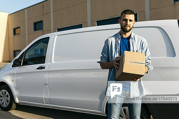Young delivery man with package standing in front of van