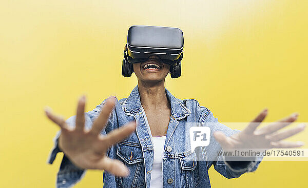 Smiling young woman wearing virtual reality headset gesturing against yellow background