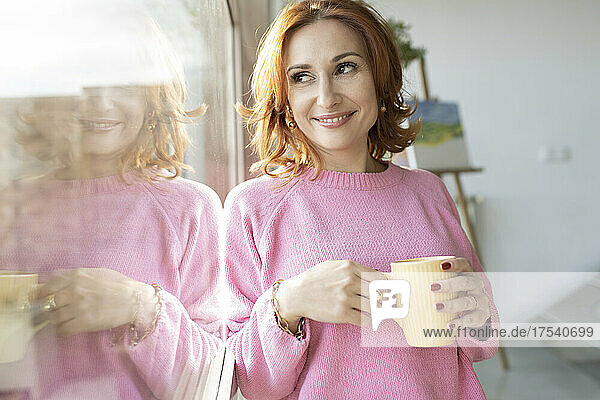 Smiling woman holding mug looking out of window at home