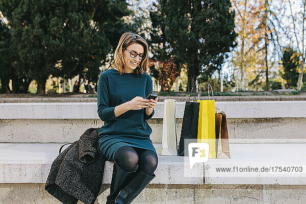 Smiling woman using smart phone sitting on steps by shopping bags