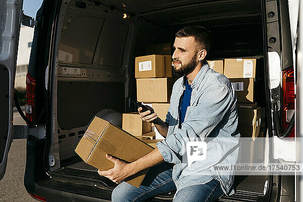 Thoughtful delivery man with package and smart phone sitting in van