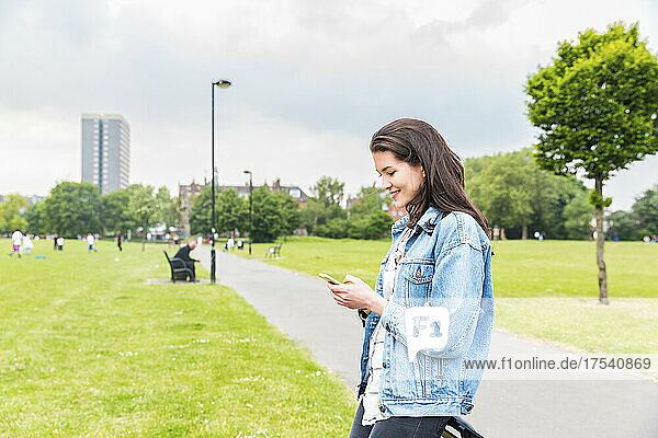 Smiling young woman wearing denim jacket text messaging on smart phone at park