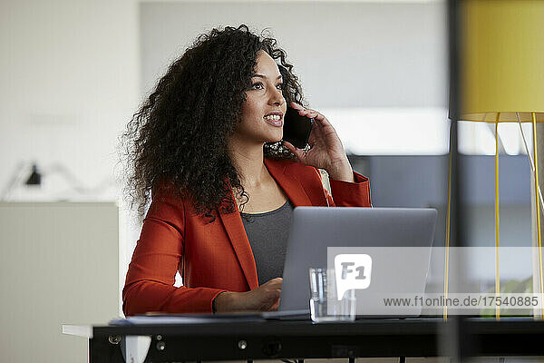 Businesswoman with laptop talking on smart phone in office