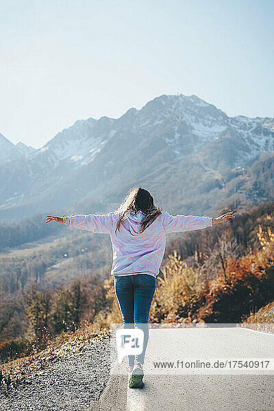 Woman with arms outstretched walking at roadside towards mountains
