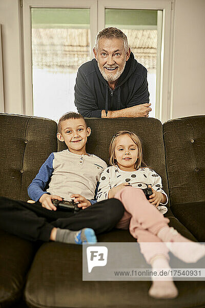 Girl and boy playing video game with grandfather leaning on sofa at home