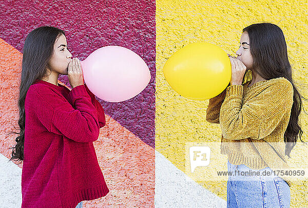 Playful sisters blowing up balloon next to multi colored wall