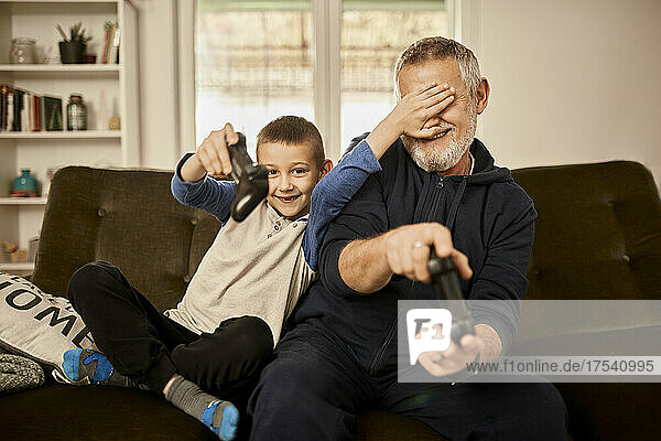 Enkel verdeckt die Augen seines Großvaters  der zu Hause ein Videospiel spielt