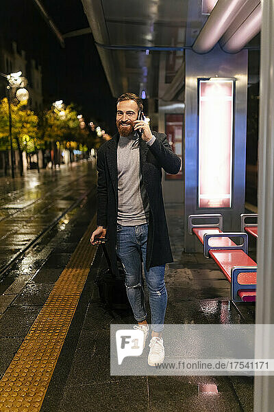 Smiling man holding suitcase and talking on mobile phone at tram station