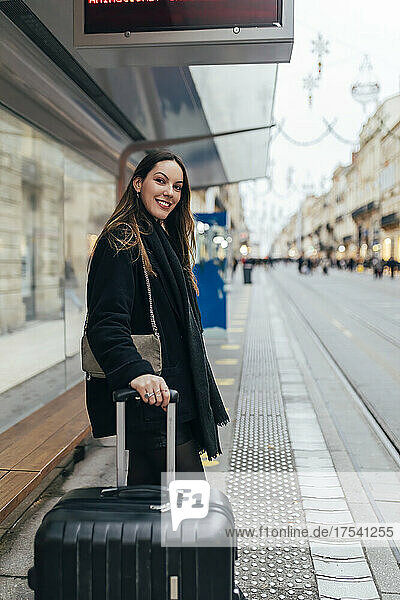 Smiling woman with suitcase waiting at tram station in city