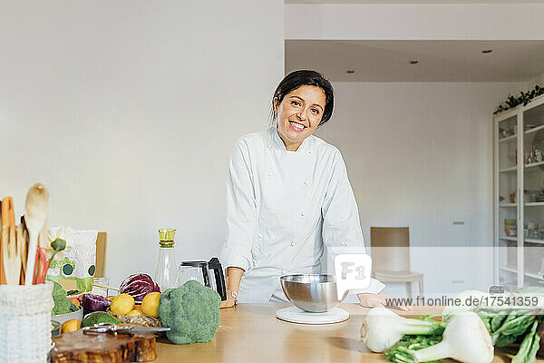 Smiling chef leaning on table with green vegetables at home