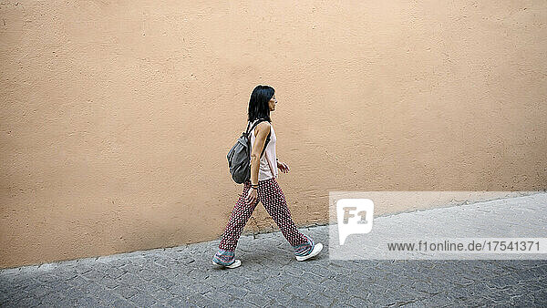 Woman with backpack waking on footpath by peach wall