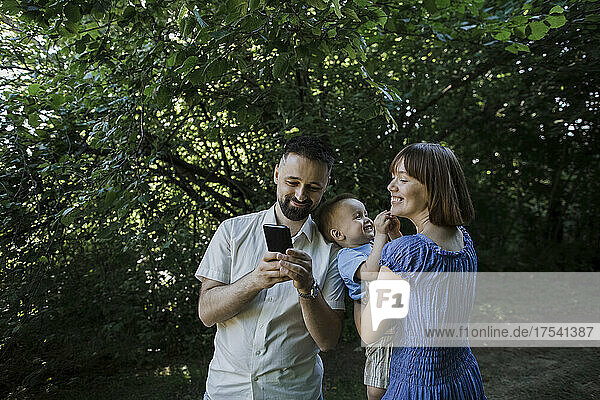 Smiling woman carrying son and looking at man showing smart phone