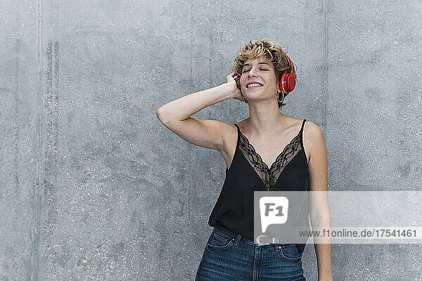 Lächelnde Frau hört Musik über Kopfhörer vor grauer Wand