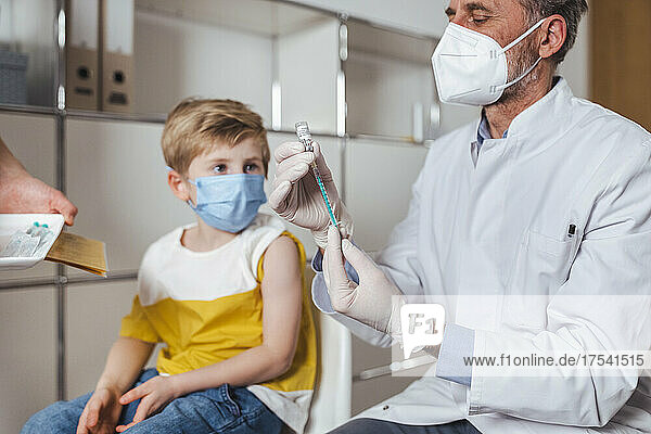 Doctor with face mask preparing vaccine injection for boy at center