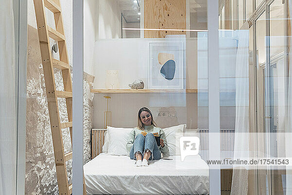 Smiling woman reading book sitting on bed at home