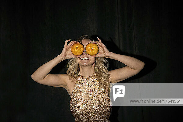 Playful woman covering eyes with oranges against black background
