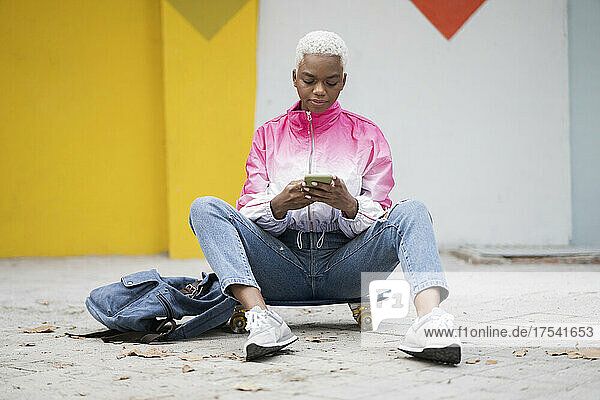 Junge Frau sitzt mit Rucksack auf Skateboard und benutzt Smartphone
