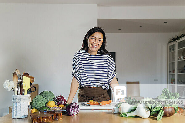 Lächelnde Frau steht mit frischem Gemüse am Tisch