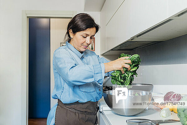 Woman taking out washed spinach from steel bowl in kitchen at home