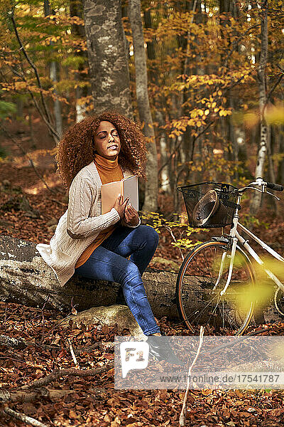 Woman with eyes closed sitting on log holding book by bicycle in autumn forest