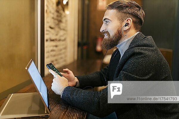 Smiling businessman holding mobile phone and listening music through in-ear headphones