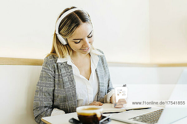 Young freelancer wearing headphones writing in diary at cafe
