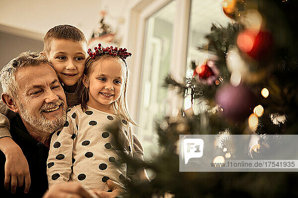 Grandchildren with grandfather looking at decorated christmas tree at home