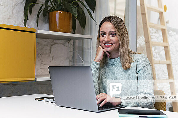 Beautiful businesswoman with hand on chin sitting with laptop at studio