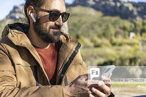 Bearded man wearing sunglasses using mobile phone and listening music through wireless in-ear headphones