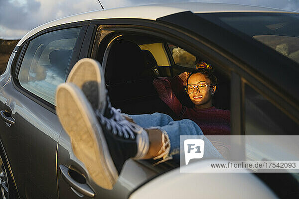 Happy woman sitting with feet up on car window at sunset