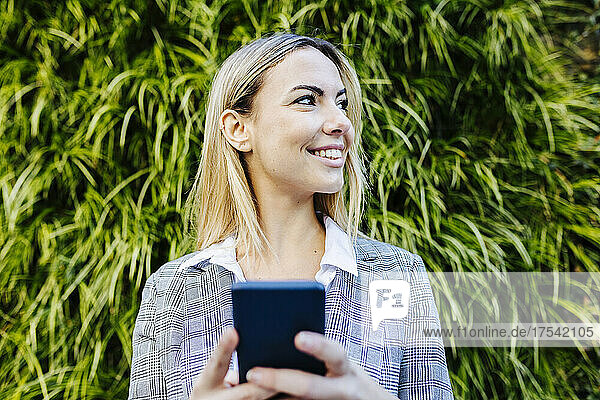 Young blond working woman with smart phone in front of green plants