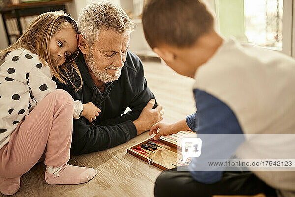 Granddaughter embracing grandfather looking at boy playing backgammon at home