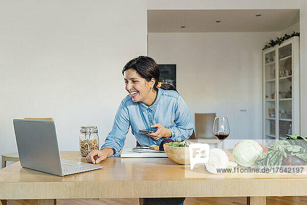 Smiling woman holding smart phone on video call using laptop at home