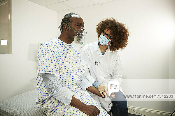 Doctor wearing protective face mask sitting with patient inhaling through nebulizer in medical room