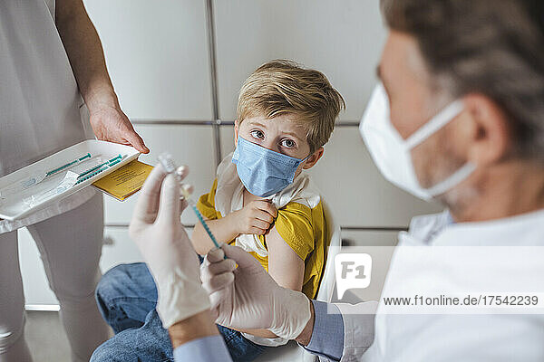 Boy with face mask looking at doctor preparing injection at vaccination center