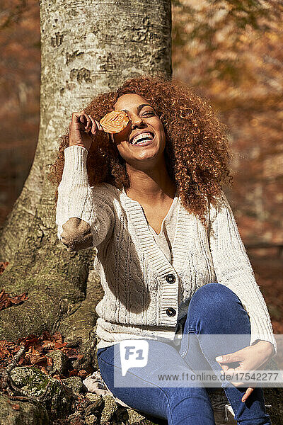 Happy woman covering eye with leaf on sunny day in autumn forest