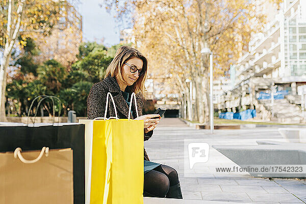 Woman using smart phone sitting by shopping bags