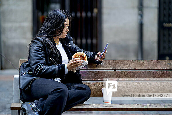 Woman with burger using mobile phone on bench