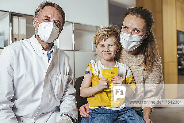 Smiling boy holding vaccination certificate with doctor and mother at center