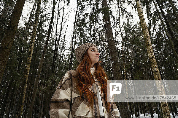 Thoughtful redhead woman wearing knit hat in winter forest