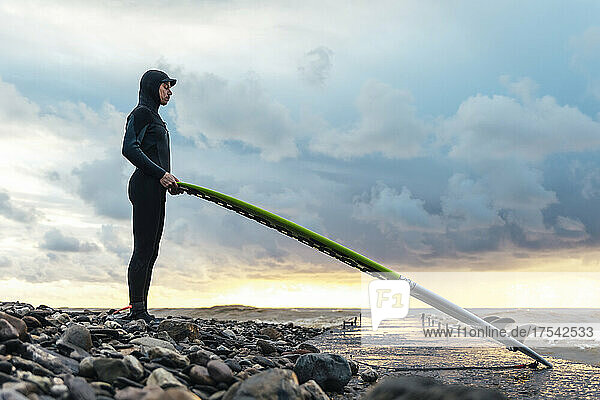 Surfer wearing wetsuit holding surfboard standing on pebbles near sea