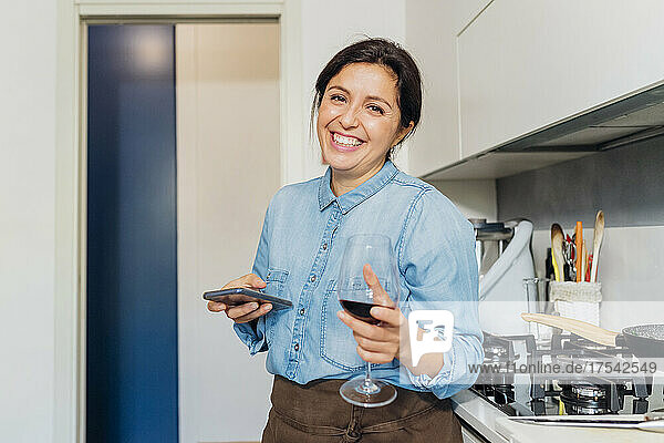 Happy woman with smart phone holding wine glass in kitchen at home