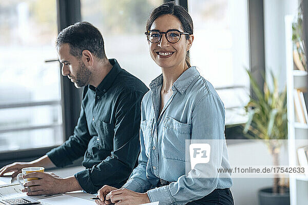 Happy architect wearing eyeglasses sitting by colleague working in office