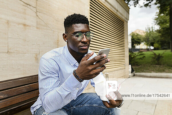 Young man with lunch box using smart phone on bench