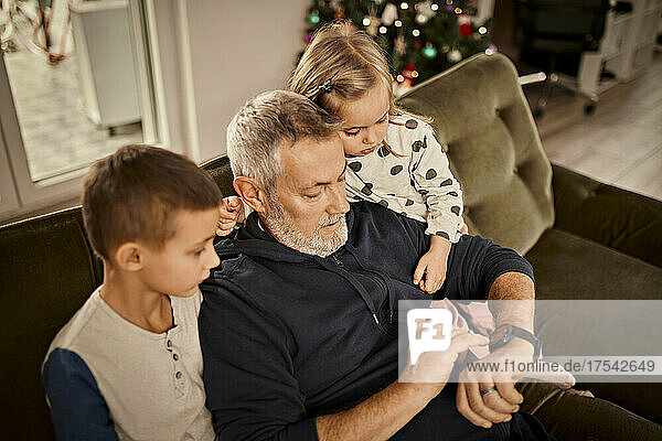 Grandfather teaching controls of smart watch to grandchildren sitting on sofa at home