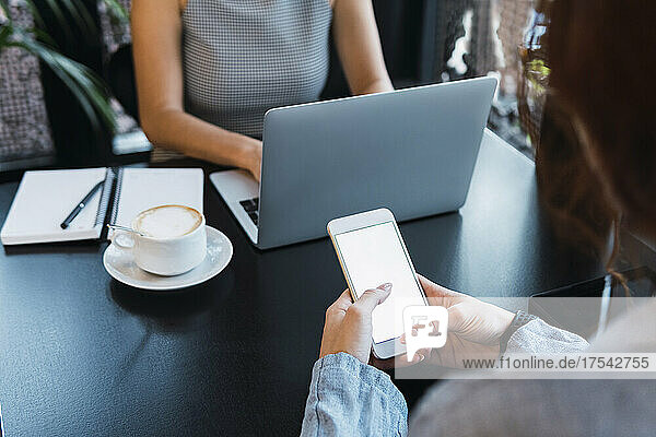 Businesswoman using smart phone by colleague working on laptop at coffee shop