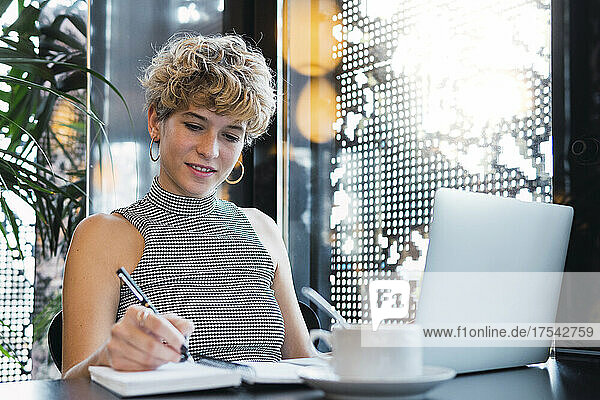 Young businesswoman with laptop writing in diary at cafe