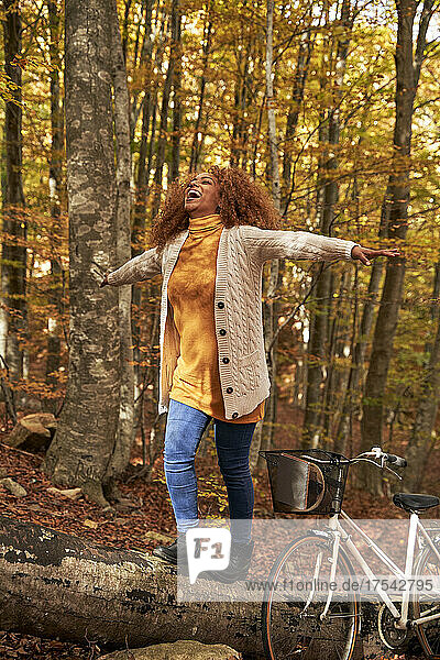 Happy woman with arms outstretched walking on log by bicycle in autumn forest